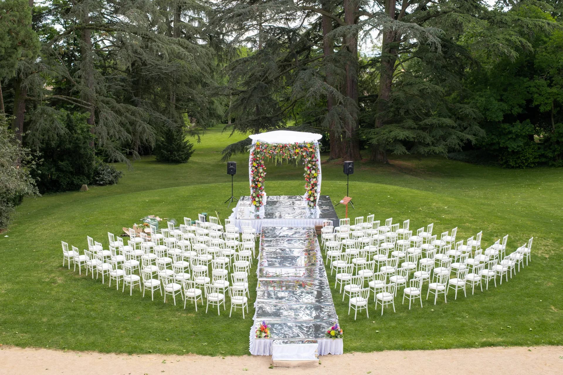 Vue d'ensemble d'une cérémonie laïque dressée dans un parc arboré : arche fleurie, allée blanche et rangées de chaises