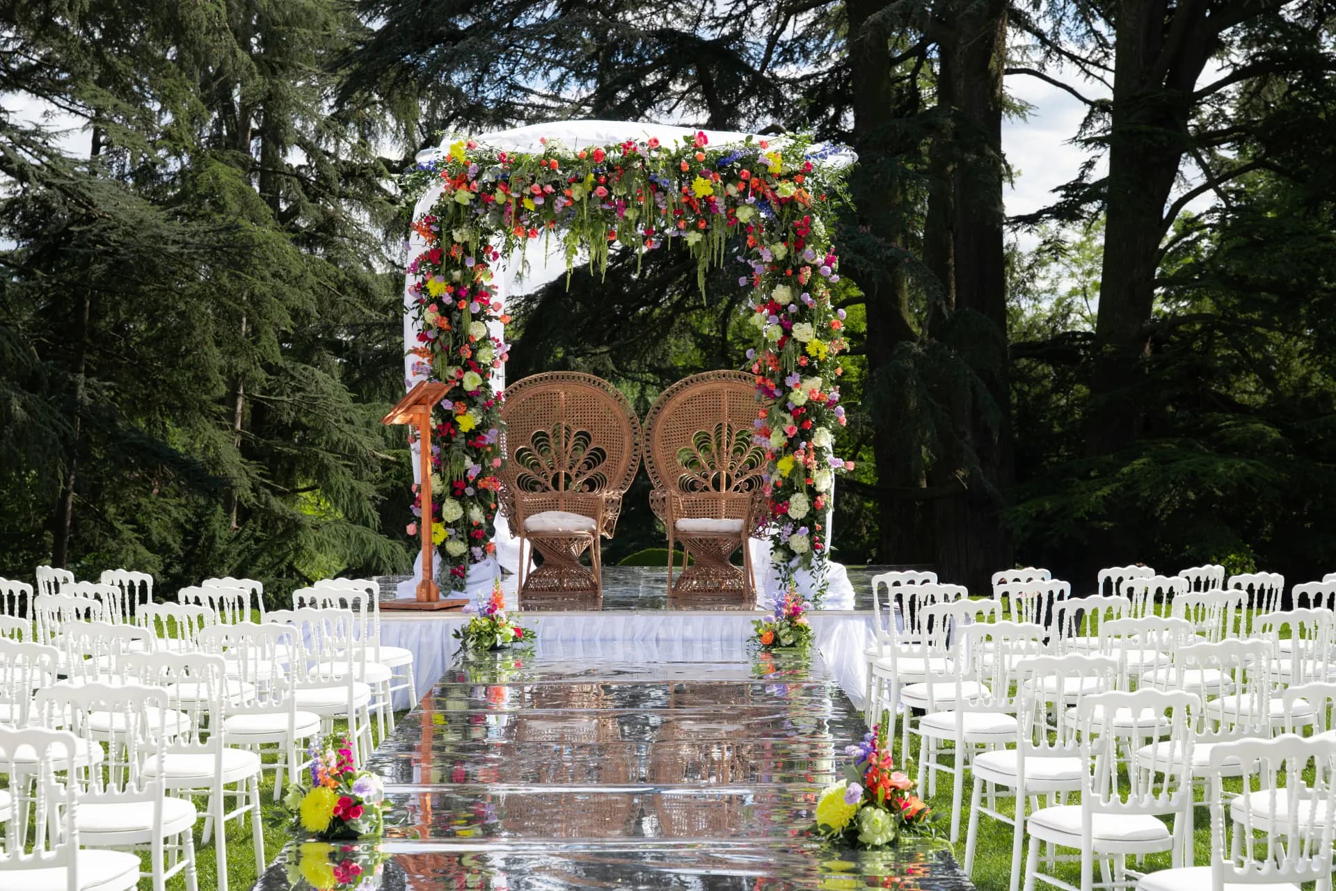 Arche de cérémonie fleurie et fauteuils en rotin au bout d'une allée miroir bordée de chaises blanches, dans un parc arboré