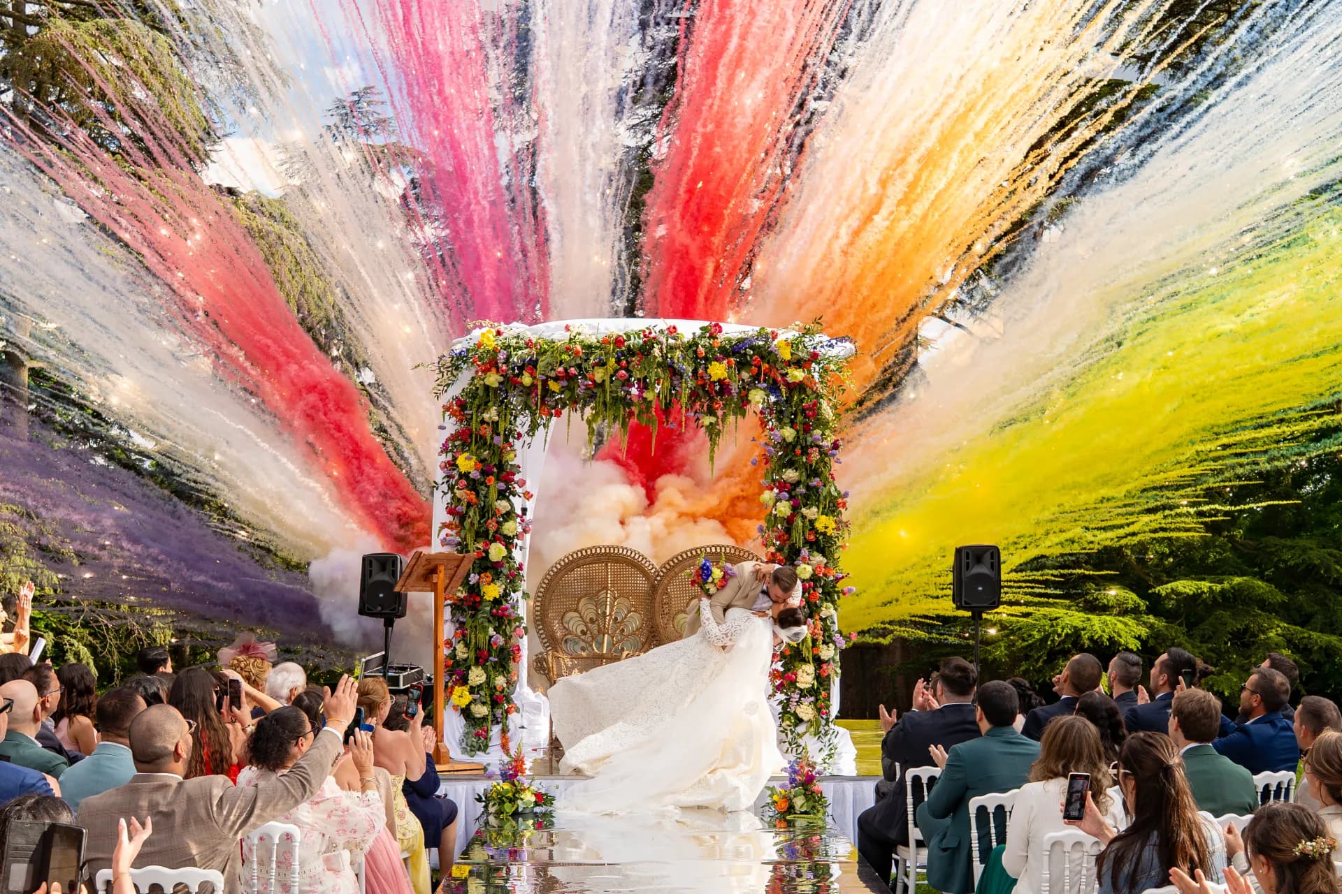 Cérémonie laïque en extérieur : les mariés s'embrassent sous l'arche fleurie tandis que les canons à confettis multicolores explosent devant les invités